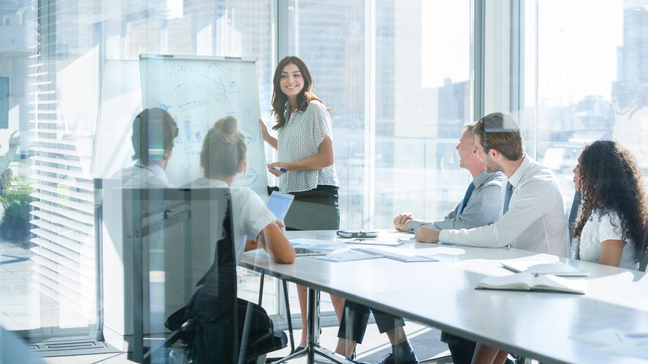 A woman stands next to a paper flip chart while presenting to a diverse group of people in business attire in a room with floor to ceiling windows